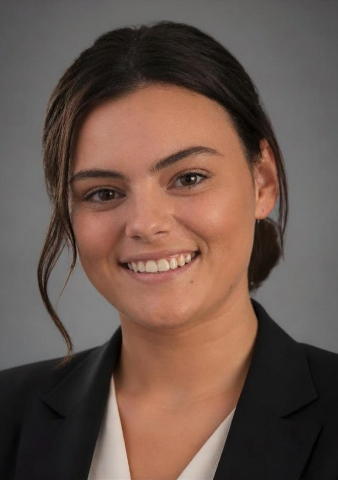 A smiling woman with dark brown hair pulled back, wearing a black blazer over a white top, photographed against a plain gray studio background.